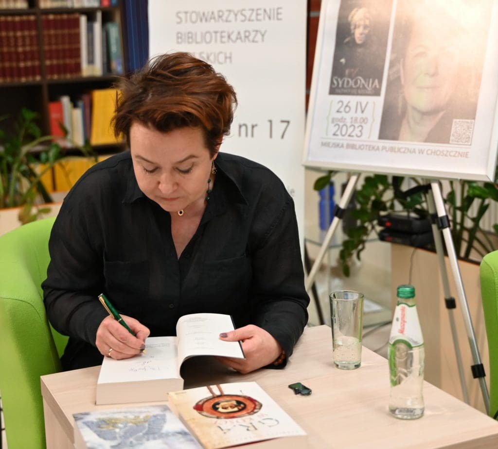 Elżbieta Cherezińska, pisarka podpisuje książkę. Na stoliku leżą książki i stoi butelka wody oraz szklanka.